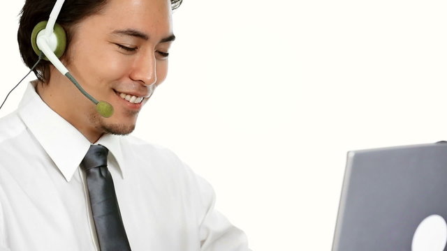 An attractive businessman wearing a white shirt and grey tie, with a headset. Talking on the phone while working on his computer. White background.