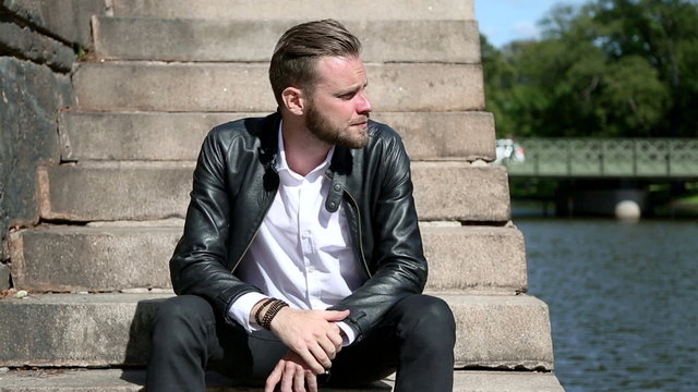 A young and attractive man in his 20s sitting down outside on a set of steps, wearing a white shirt, black jeans, with a black leather jacket. A sunny summer day.
