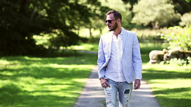 Man in his 20s walking along a path with greens grass and trees around him, a sunny summer day. Wearing a light blue blazer and jeans with sunglasses. 