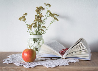Vintage still life with an open book, glasses, apple and vase with flowers on doily