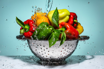 Peppers of different colors in a colander under running water.