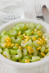 salad celery with corn in white bowl and glass of water on white wooden background