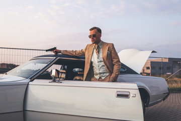 Retro 1970s gangster with pistol leaning against vintage car.