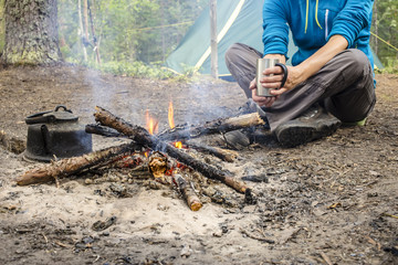 Girl in a blue jacket sitting while camping near the bonfire close to tent is heated and drink hot tea