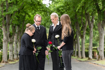 Familie bei Beerdigung trauert auf Friedhof, steht in Gruppe mit Blumen auf einer Allee