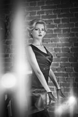 Young beautiful short hair blonde woman in black dress posing, black and white photo. Elegant romantic mysterious lady with movie star look, bricks wall on background. Retro style blonde with gloves