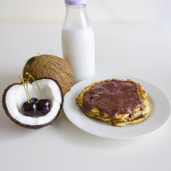 Breakfast and brunch coconut pancakes meal with cracked open coco, glass bottle of milk and cherry on white background. Stack of crepes is topped with cream syrup ready to eat.