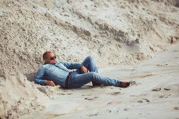 gorgeous young man in glasses lies in jeans clothes on the sand
