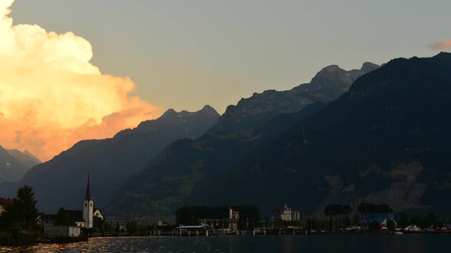 Evening clouds are painted in bright orange color. Mountain landscape.Reflections in the lake. In the distance the small town of in Fluelen in Switzerland. Time Lapse