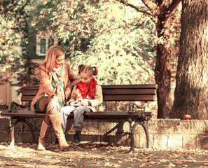 Mother and daughter sitting on a bench in the park and playing with doll.