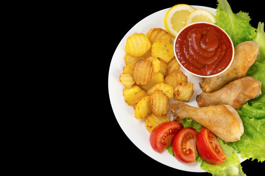 chicken legs on a white plate with slices of tomato and lettuce and french fries and ketchup top view isolated on black background
