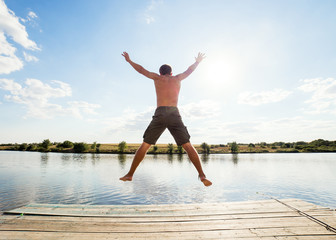 Fototapeta na wymiar Happy man jumping on pier with lake and sky in background