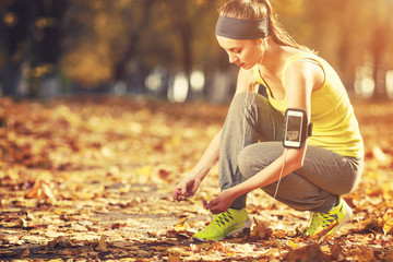 Running woman. Runner is tying laces and listening to music. Female fitness model tying laces outside in autumn park. Woman athlete training in the fall outdoors background. Sport lifestyle.
