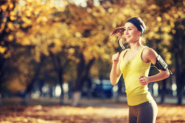 Female fitness model training outside on a warm fall day and listening to music using smart phone. Young woman jogging in autumn park. Sport lifestyle.
