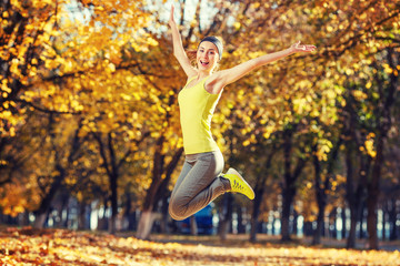 Young happy woman jumping in autumn park. Female fitness model training outside on a warm fall day....