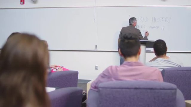 A professor finishes up writing on the whiteboard and addresses the class
