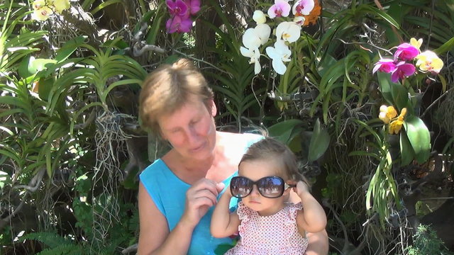 baby sitting on the lap of a woman in a blue dress and playing with sunglasses on the background of trees and beautiful multi-colored orchids. the child takes, and then puts sunglasses
