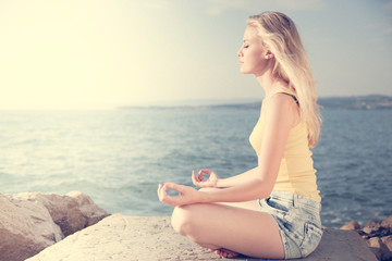 Beautiful young blond woman meditating on a beach at sunrise in