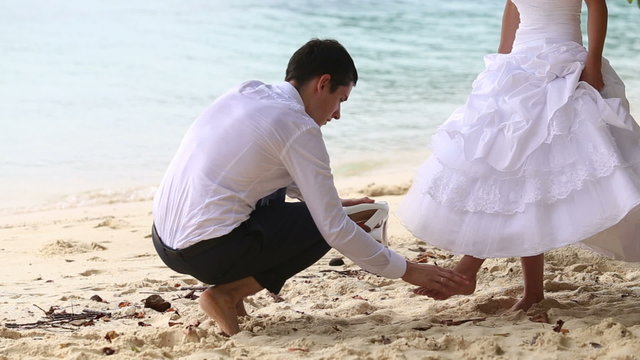 groom puts shoe on long haired bride under tree on beach