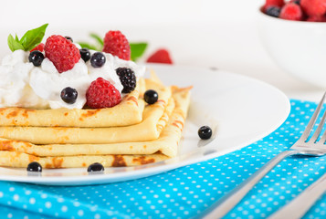 Delicious sweet crepes decorated air-cream and ripe berries, raspberries, blackberries and blueberries on a white plate, bright blue polka dot napkin. Tasty breakfast.