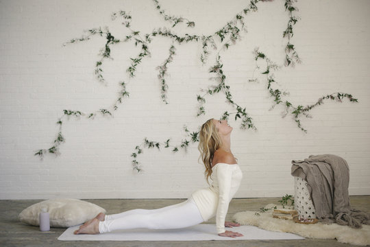 A blonde woman in a white leotard and leggings, lying on a white mat in a room, arching he back in a stretch.