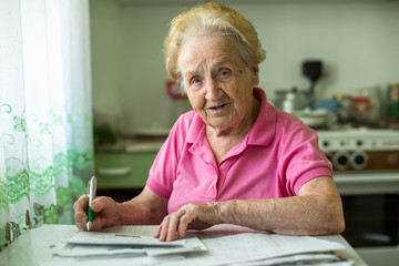 Senior woman populates handle her utility bills notices, sitting at the table in the kitchen.