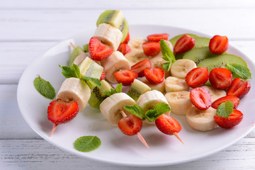 Fresh fruits on skewers in plate on wooden table, closeup