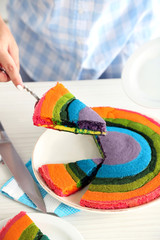 Young woman cutting rainbow cake in kitchen