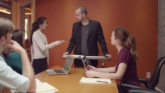 Man leading a small business meeting when an intern brings him a note and he leaves