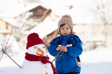 Happy beautiful child building snowman in garden, winter