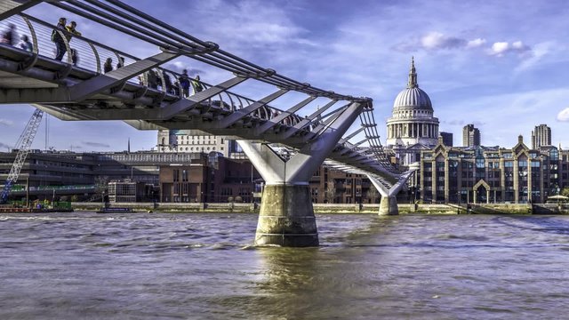 Timelapse view of the Millennium bridge in London and St Paul cathedral, with pedestrians, tourists and commuters walking and boats crossing the river Thames