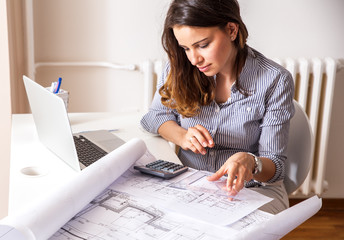Female architect working at home.She looking at blueprint. - obrazy, fototapety, plakaty