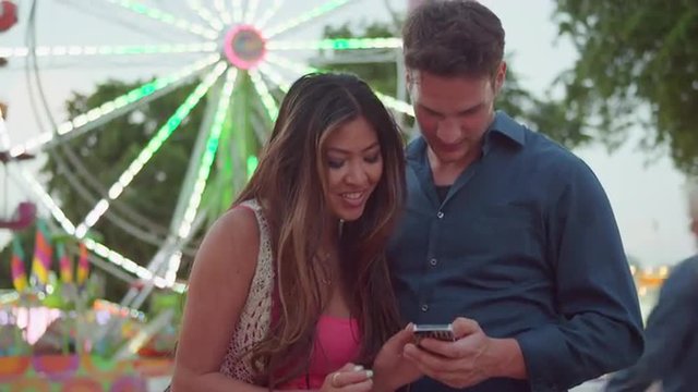 A couple taking selfies in front of a ferris wheel and looking at the pictures
