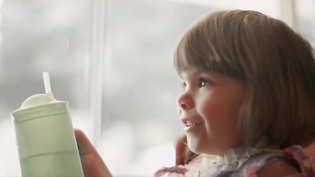 A little girl wearing a princess dress sits by a window and drinks from a sippy cup
