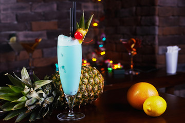 tropical blue curacao alcoholic cocktail garnished with a fresh pineapple slice and maraschino cherries on a table in the restaurant with bright backgrounds of beautiful disco lights.  - obrazy, fototapety, plakaty