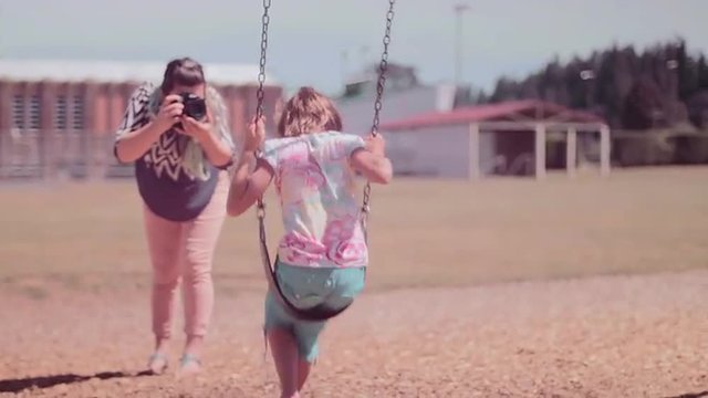 A mother taking pictures of her daughter on a swingset

