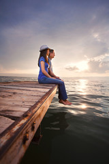 Fototapeta premium Side view of young couple sitting on the pier watching sunset or sunrise