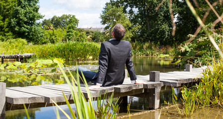 corporate green break - relaxed middle age male professional sitting on a wooden bridge in the middle of an asian-like green pond for company wellbeing,back view with park foreground