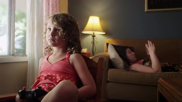 A little girl in a chair controls the TV while her mother uses her phone on the couch
