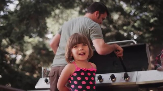 A little girl watches her dad clean a barbecue
