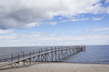 horizontal image of a long wooden pier jutting out over the ocean into the distance under a blue summer sun with clouds floating by with room for text.