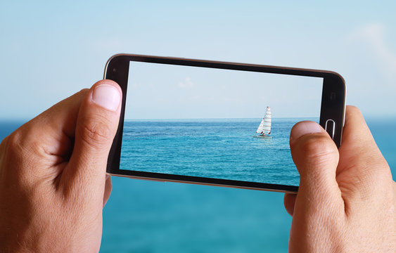 Male hand taking photo of Sailboat, sea, sun, blue, sea, horizon and ocean with cell, mobile phone.