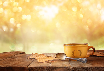 front image of coffee cup over wooden table and autumn leaves in front of autumnal sunset 