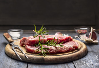 raw meat with rosemary, garlic, salt and pepper on a wooden board with a for, top view