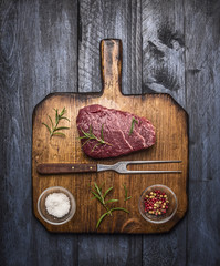 raw ribeye steak on a cutting board with a fork, with rosemary salt and pepper on rustic wooden background, top view