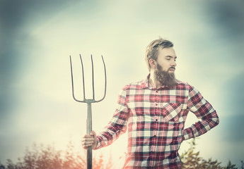 young bearded farmer in red checkered shirt with old pitchfork on sky  nature backgrund, toned
