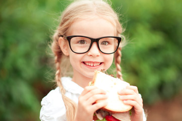 Smiling kid girl eating sandwich outdoors. Wearing glasses. Looking at camera. Childhood. Healthy...