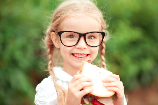 Smiling Kid Girl Eating Sandwich Outdoors. Wearing Glasses. Looking At Camera. Childhood. Healthy Lifestyle.