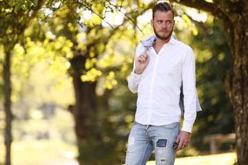 Obraz premium A man standing on green grass with trees behind him, wearing a white shirt and jeans, and holding a blazer on his shoulder. A sunny summer day.