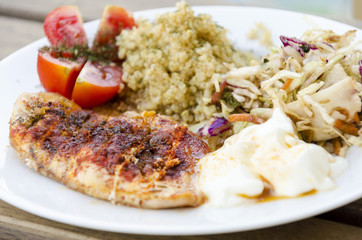 Top view of a roasted in the oven chicken breast fillet garnished with paprika served in a plate with cabbage and carrot salad, quinoa, yogurt and tomatoes on wooden table. A healthy meal.